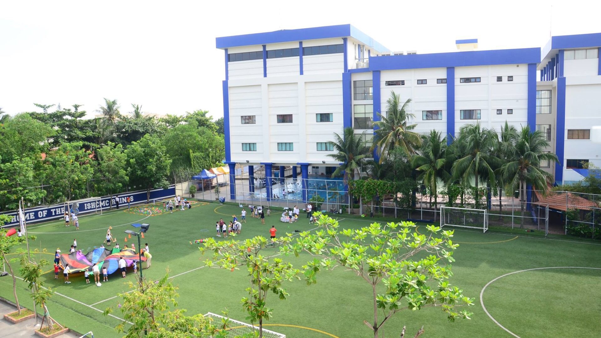 Sports field and blue-and-white classroom block at the ISHCMC campus, with a Home of the ISHCMC Lions banner